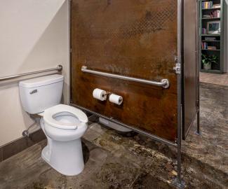Interior of an ADA toilet stall featuring a pressure-assisted toilet with grab bars, with an industrial design aesthetic.